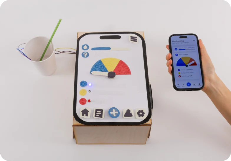 A cardboard prototype of an oversized phone with a hydration dial, next to a real phone showing the same dial and a cup with a straw.