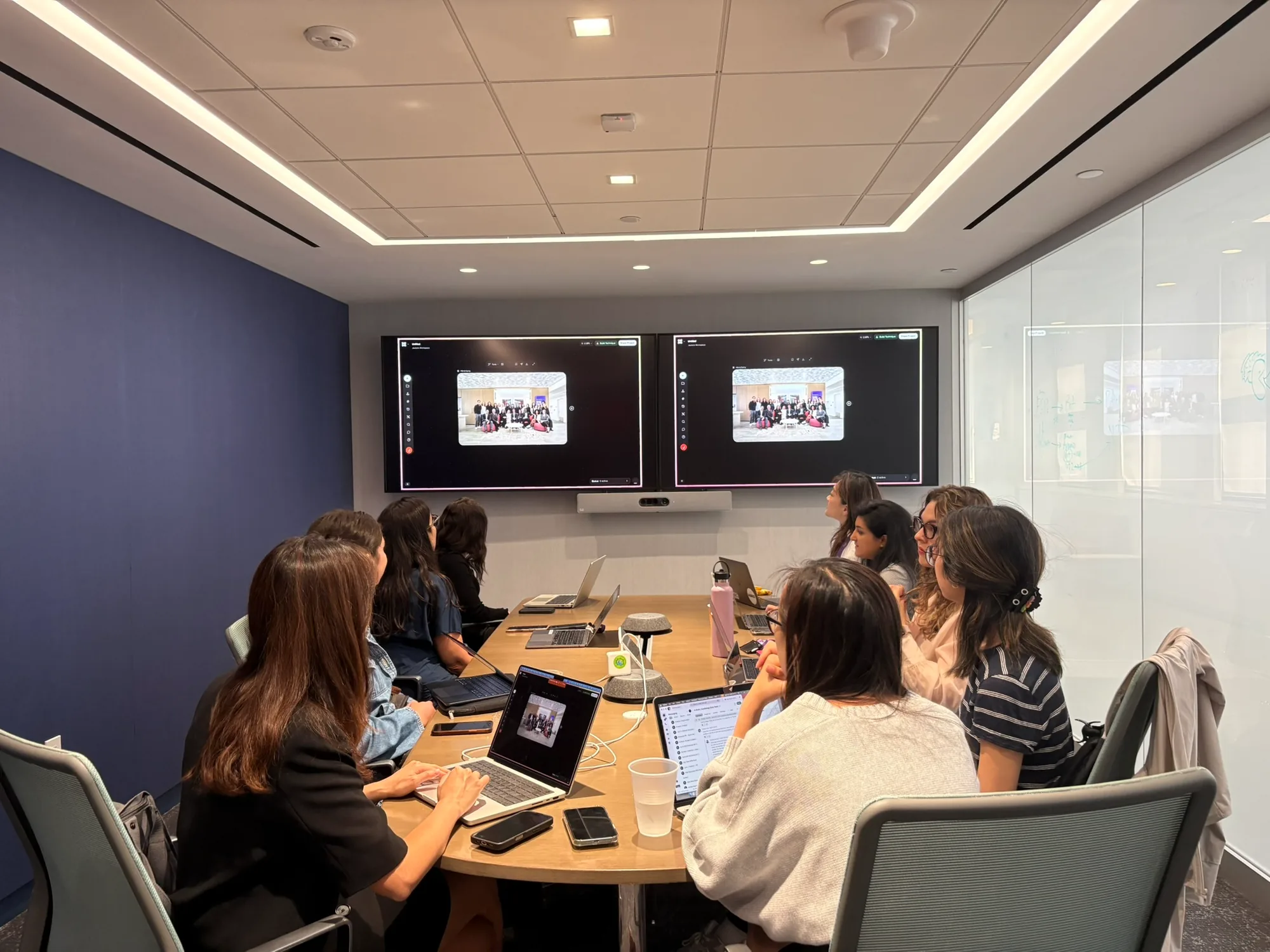 Jackie, at the bottom left, presenting from her laptop to a conference room of colleagues watching two wall screens.